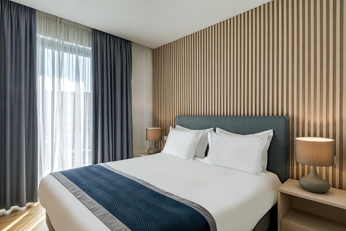 A hotel room with a large bed, two bedside tables, a headboard, and a window with curtains.