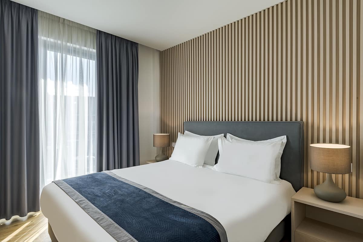 A hotel room with a large bed, two bedside lamps, a wooden slat wall, and a view of the outdoors through sheer curtains.