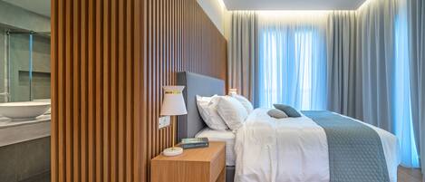 A modern hotel room with a wooden headboard, a large bed with white bedding, a bedside table with a lamp, and a view of the bathroom.