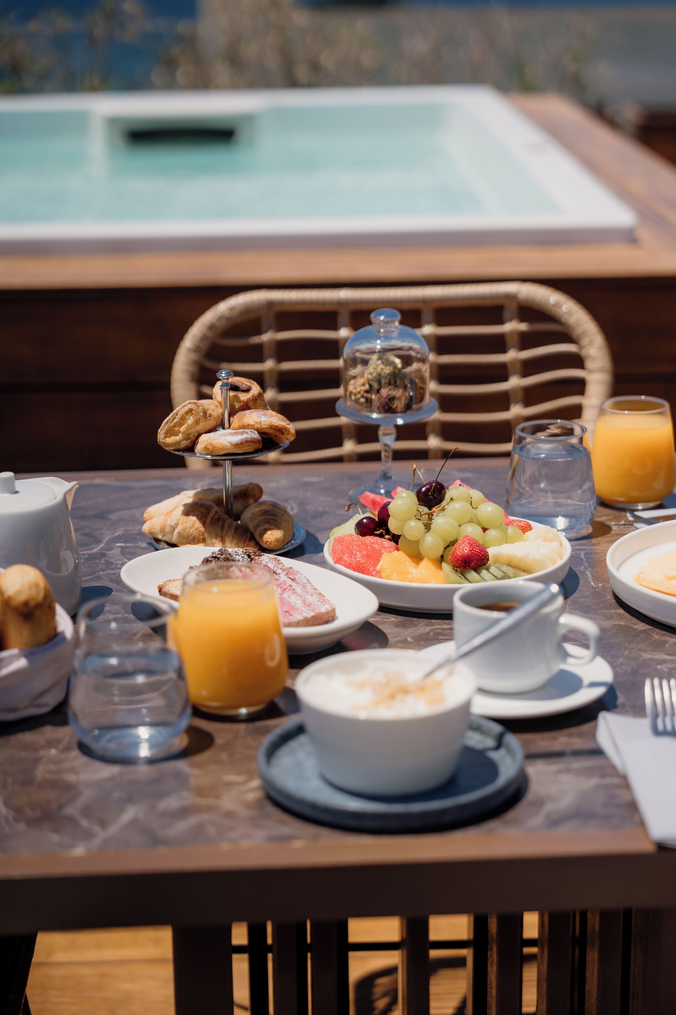 A breakfast table with various food items, a glass of orange juice, and a view of a pool in the background.
