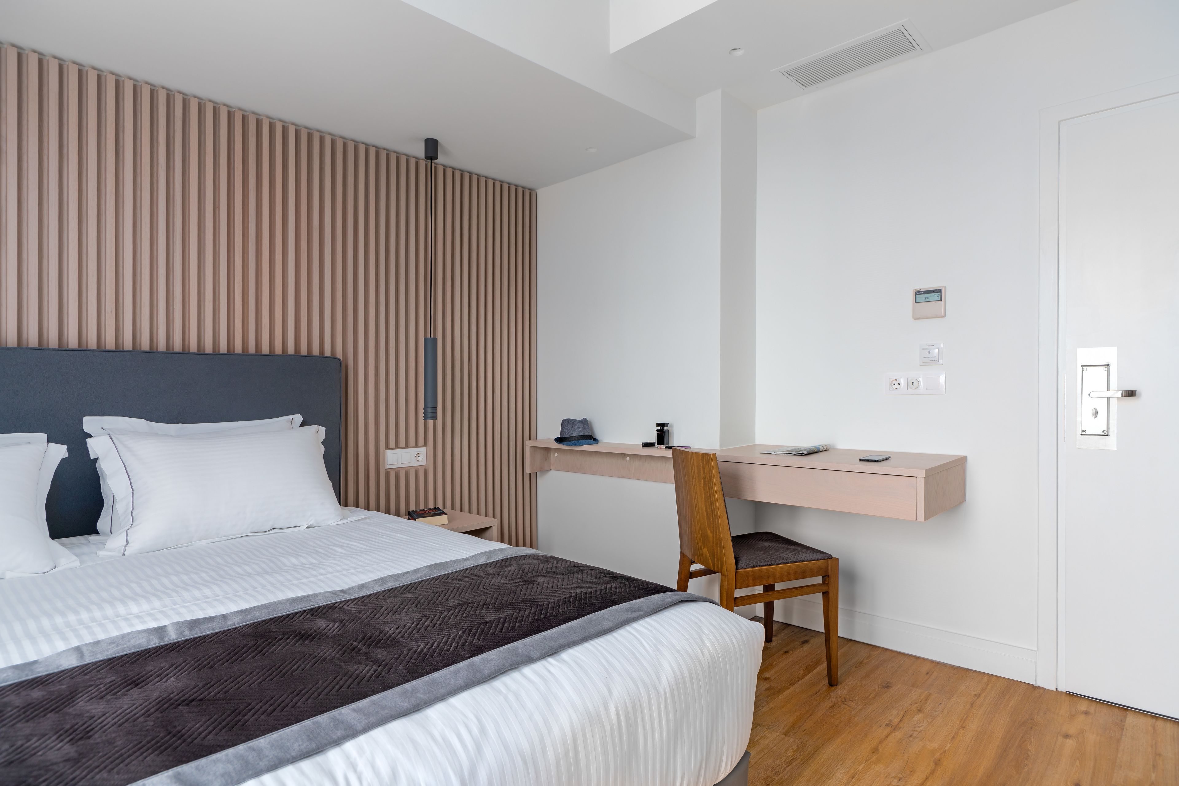 A modern hotel room with a large bed, a wooden slat wall, a desk with a chair, and a white door.