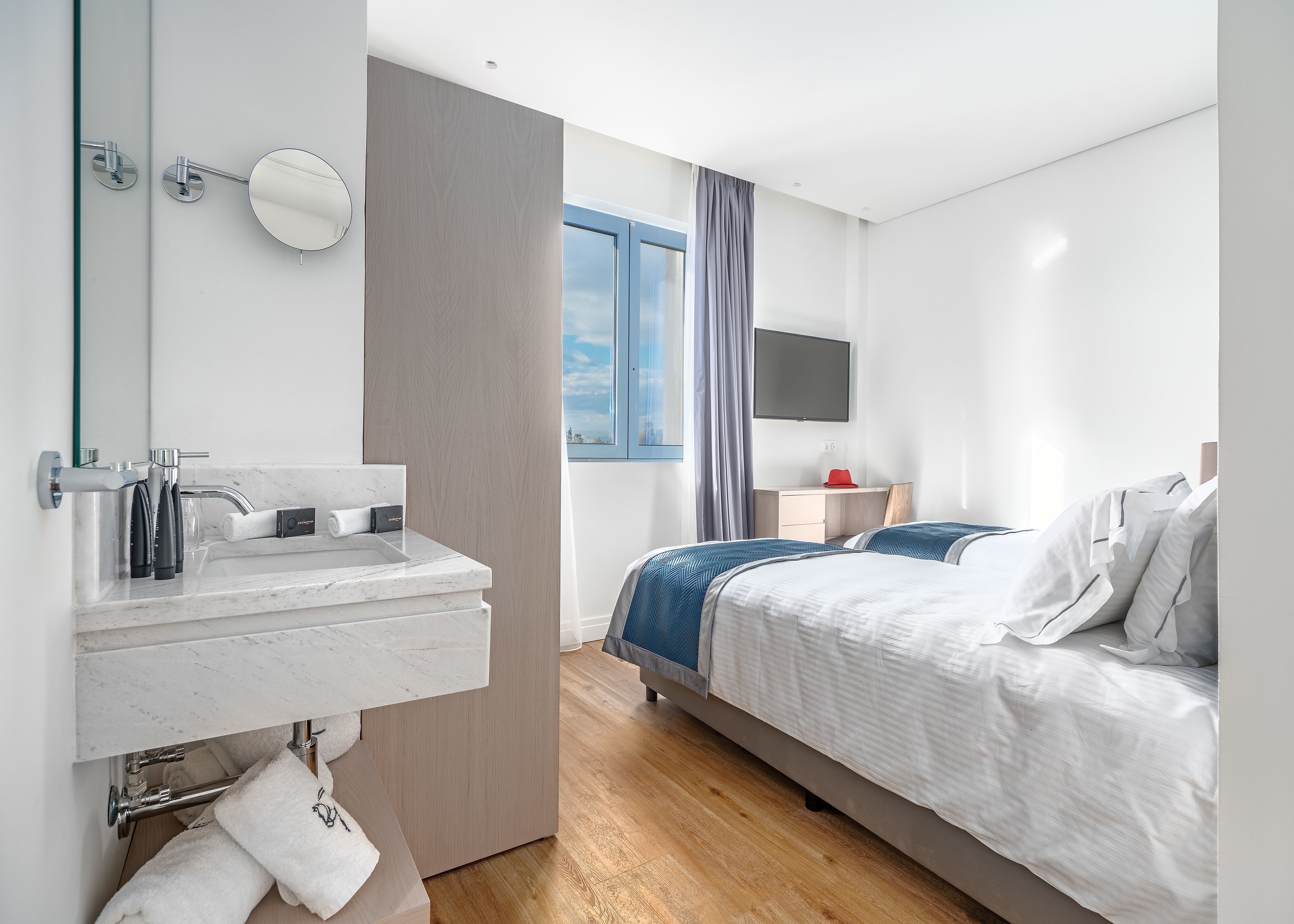A modern hotel room with a marble bathroom sink, a bed with white and blue bedding, a desk with a flat-screen TV, and a window with curtains.