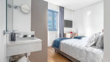 A modern hotel room with a marble bathroom sink, a bed with white and blue bedding, a desk with a flat-screen TV, and a window with curtains.
