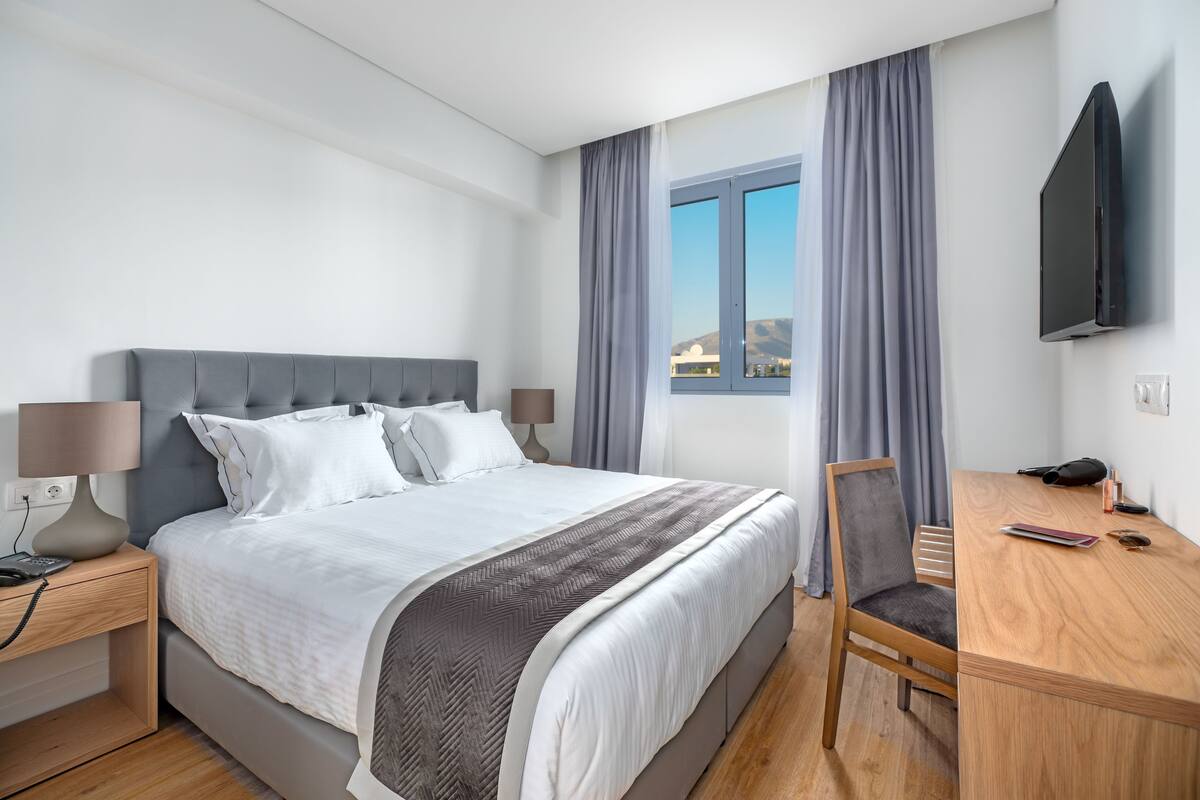 A modern hotel room with a bed, bedside table, lamp, TV, and a desk with a chair.