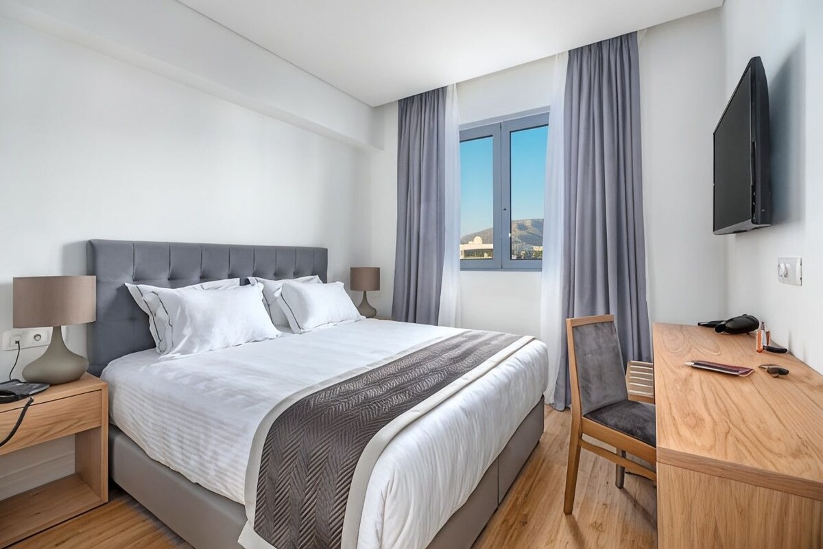 A modern hotel room with a bed, bedside table, lamp, TV, and a desk with chair.
