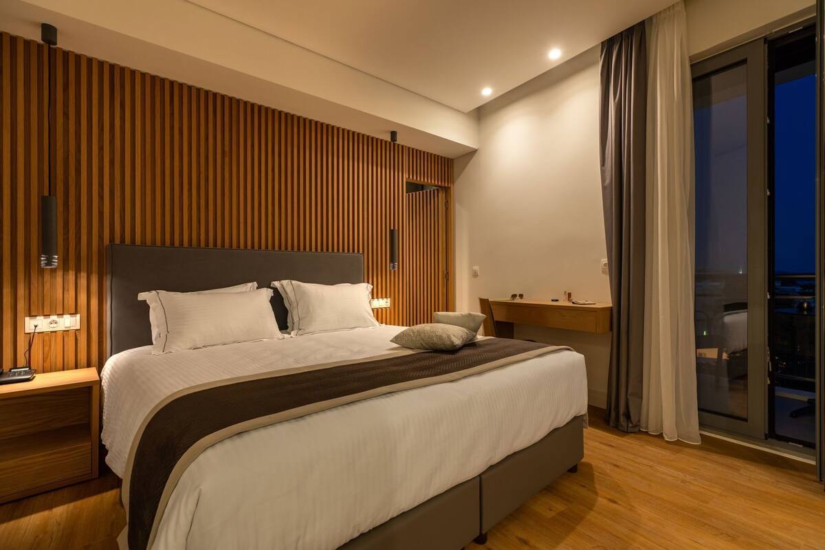 A modern bedroom with a large bed, wooden panel wall, bedside table, and a view of the city at night.
