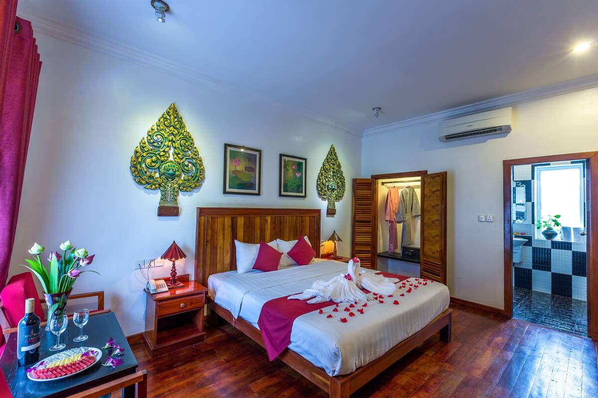 A hotel room with a wooden bed, a nightstand with a lamp, a table with a bottle and glasses, and a wall with decorative elements.