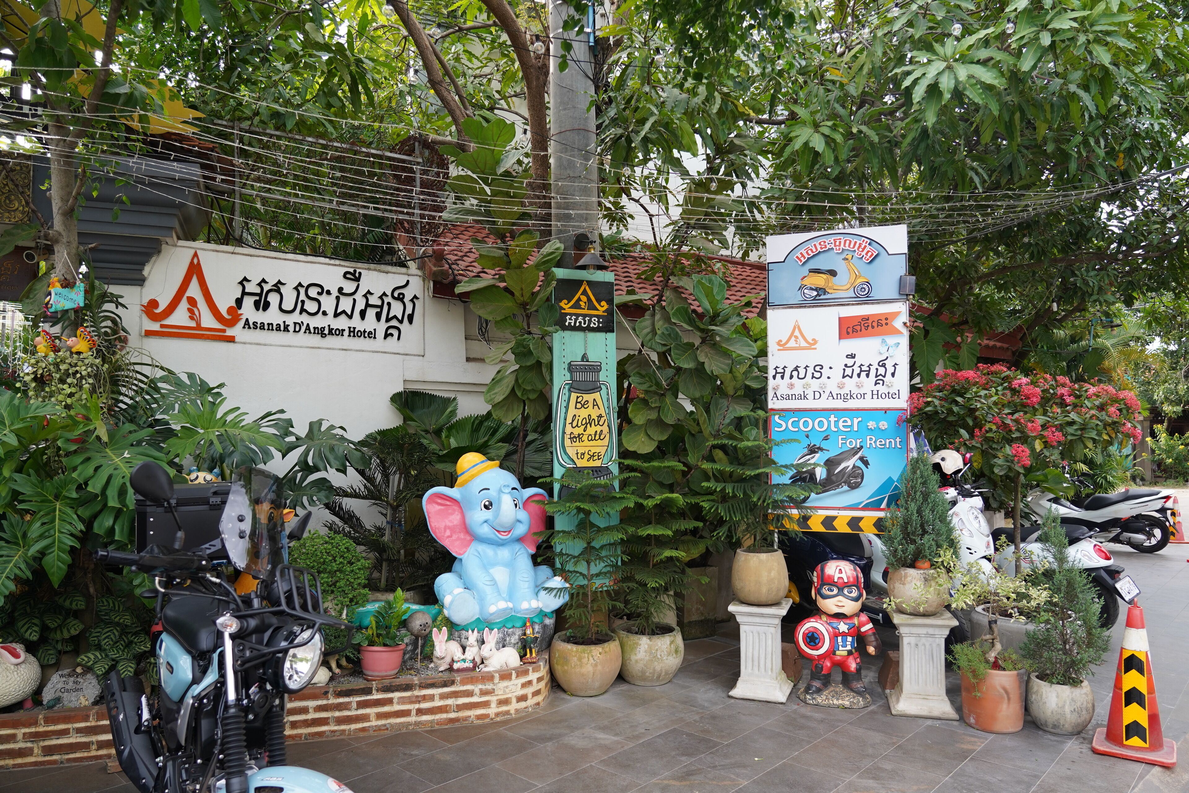 A hotel entrance with a sign for Asanak D'Angkor Hotel, surrounded by potted plants and a scooter rental sign.
