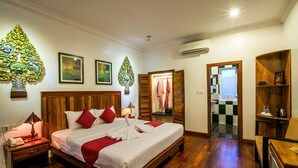 A hotel room with a wooden bed, red pillows, a wooden nightstand with a lamp, a wooden chair, and a wooden shelf with a picture frame.