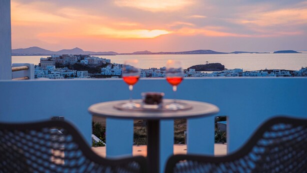 Una mesa con dos copas de vino tinto, con vistas a un pueblo costero de edificios blancos y un atardecer.