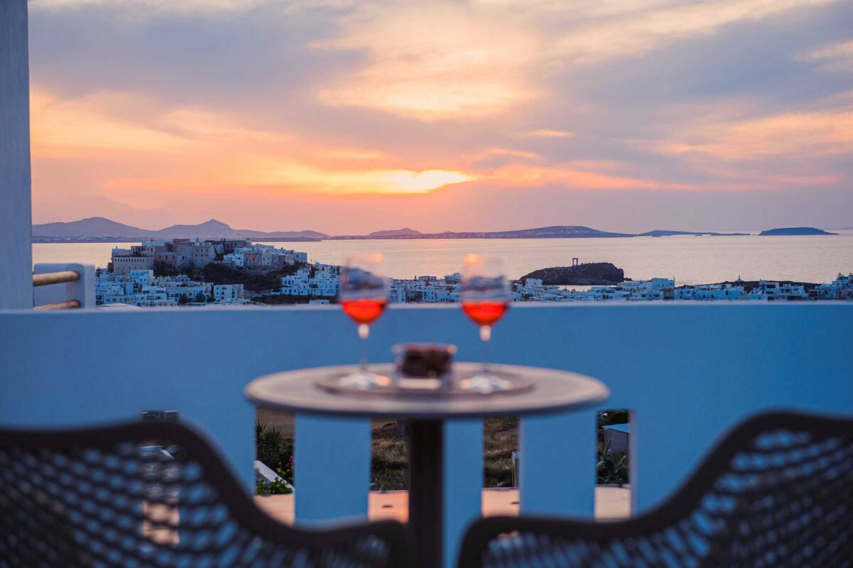 Una mesa con dos copas de vino tinto, con vistas a un pueblo costero de edificios blancos y un atardecer.