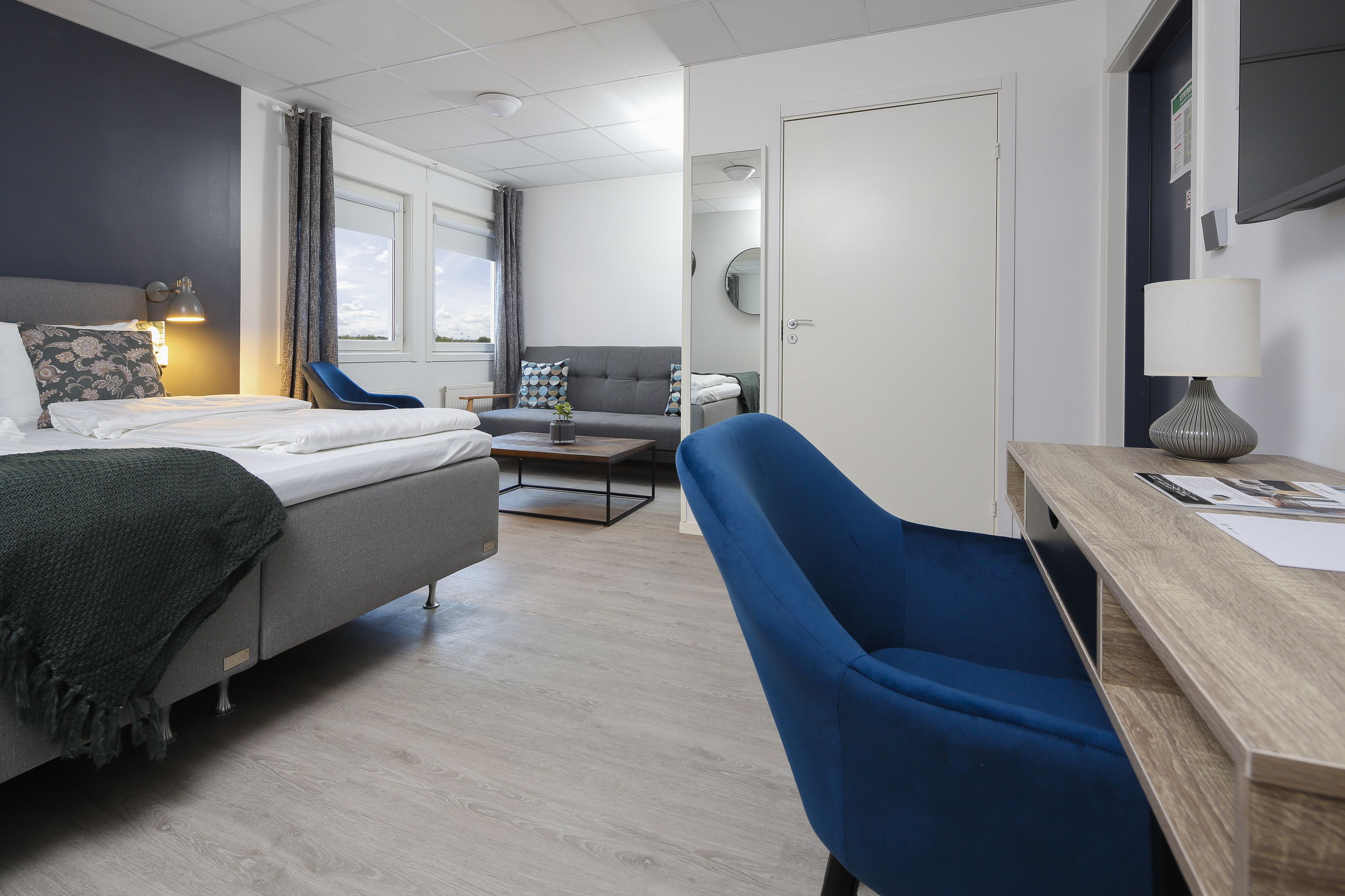 A modern hotel room with a bed, a desk, a blue armchair, and a sofa.