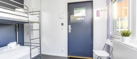 A dormitory room with bunk beds, a door with a notice, a radiator, and a window with a plant.