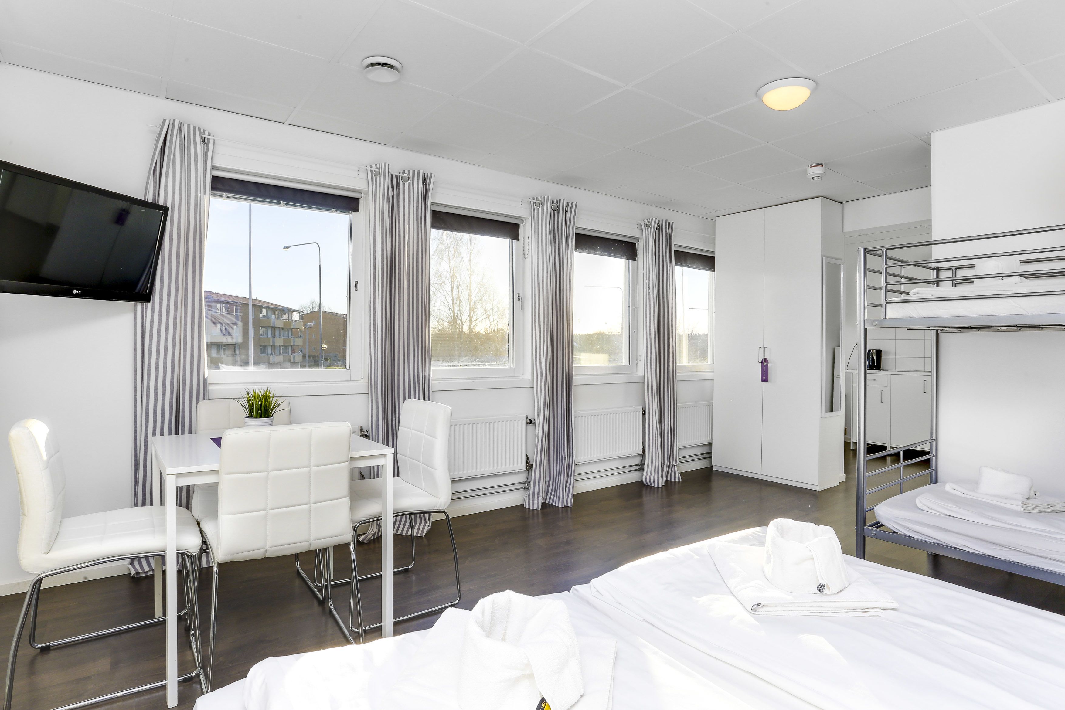 Ett hotellrum med en våningssäng, ett skrivbord med en stol, en tv som är monterad på väggen och stora fönster med gardiner.