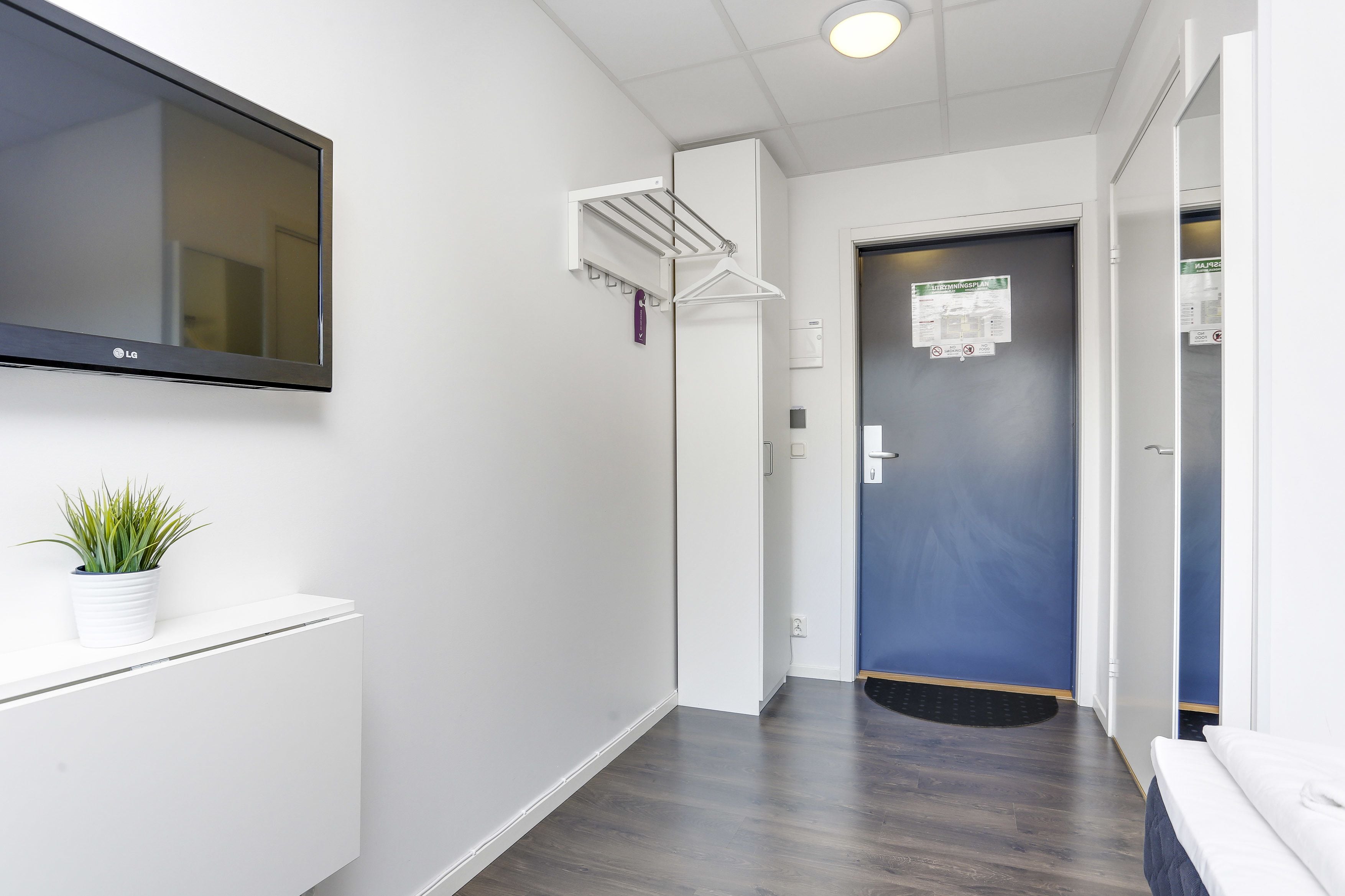 Un couloir avec un téléviseur fixé au mur, un étendoir, une porte sur laquelle est affichée une note et une plante en pot.