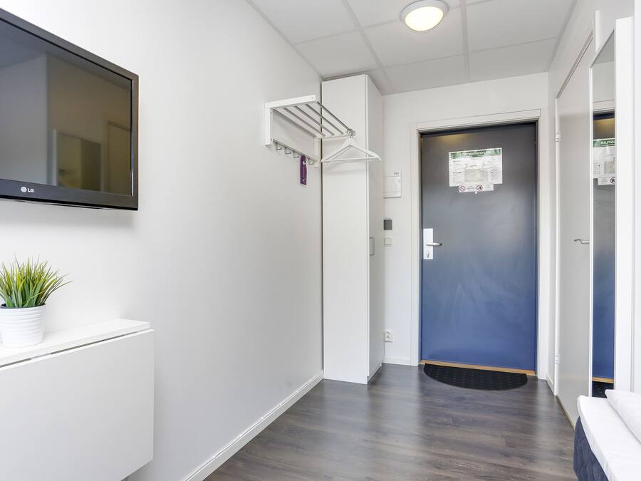 Un couloir avec un téléviseur fixé au mur, un étendoir, une porte sur laquelle est affichée une note et une plante en pot.
