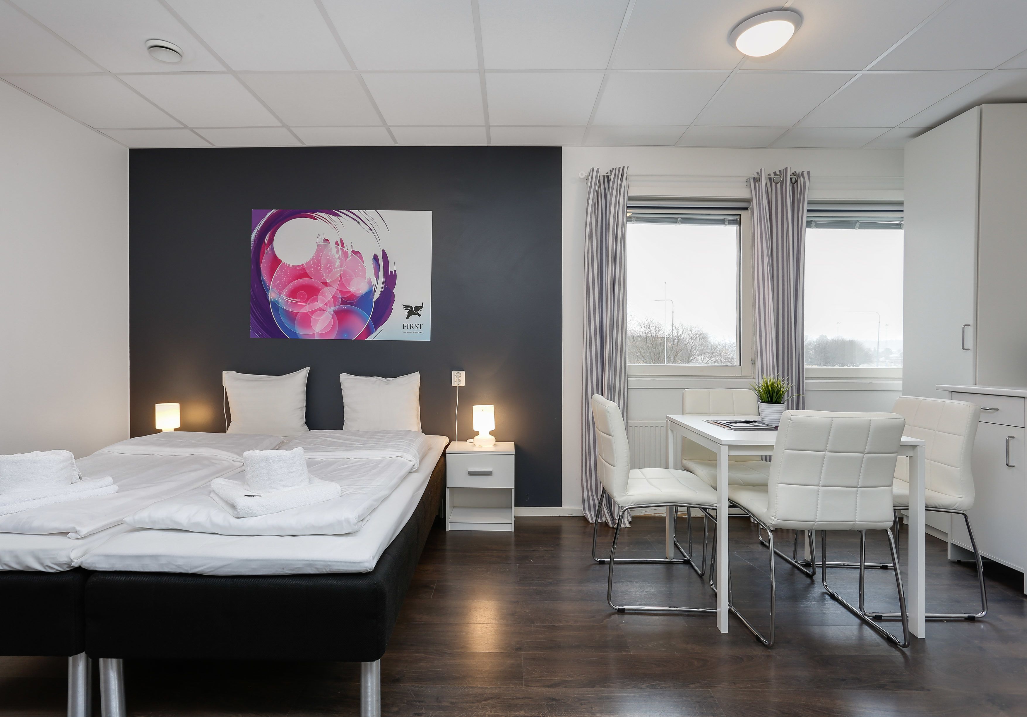 Ett modernt hotellrum med en säng, ett nattduksbord, en lampa och ett skrivbord med stolar.
