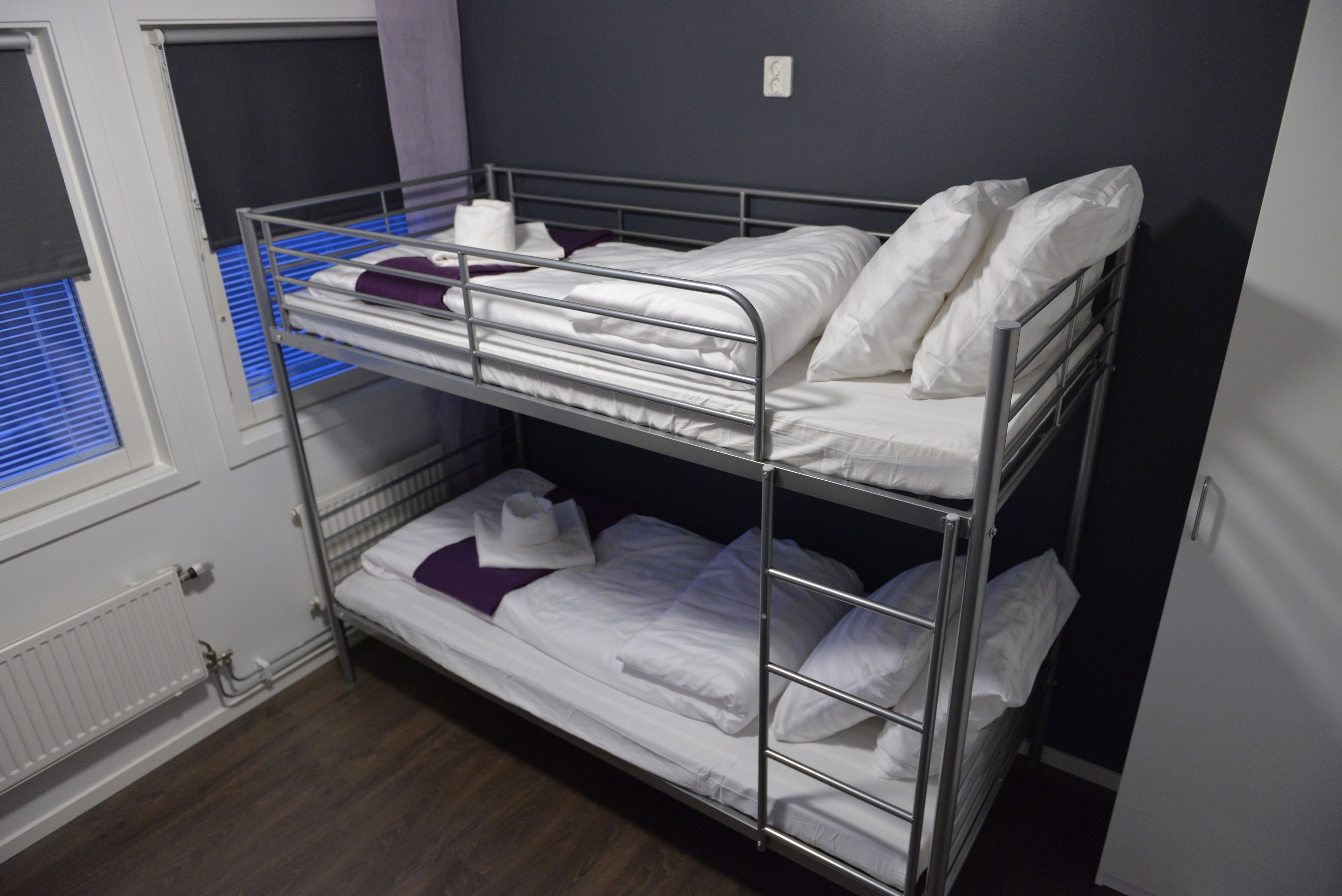 Habitación con literas, ropa de cama blanca, una ventana con persianas y un radiador.