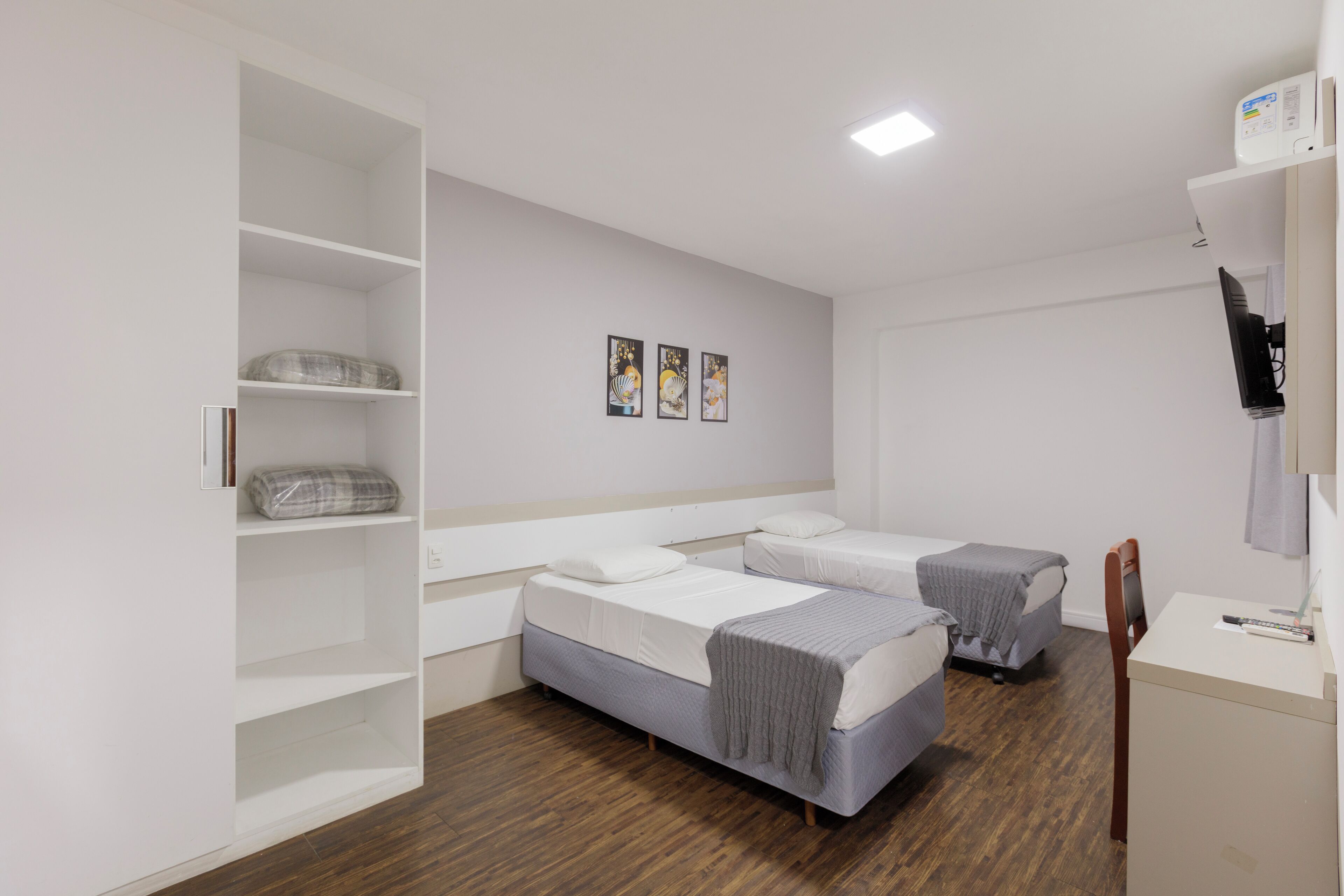A modern hotel room with two single beds, a wooden floor, a white wardrobe, and a desk with a chair.