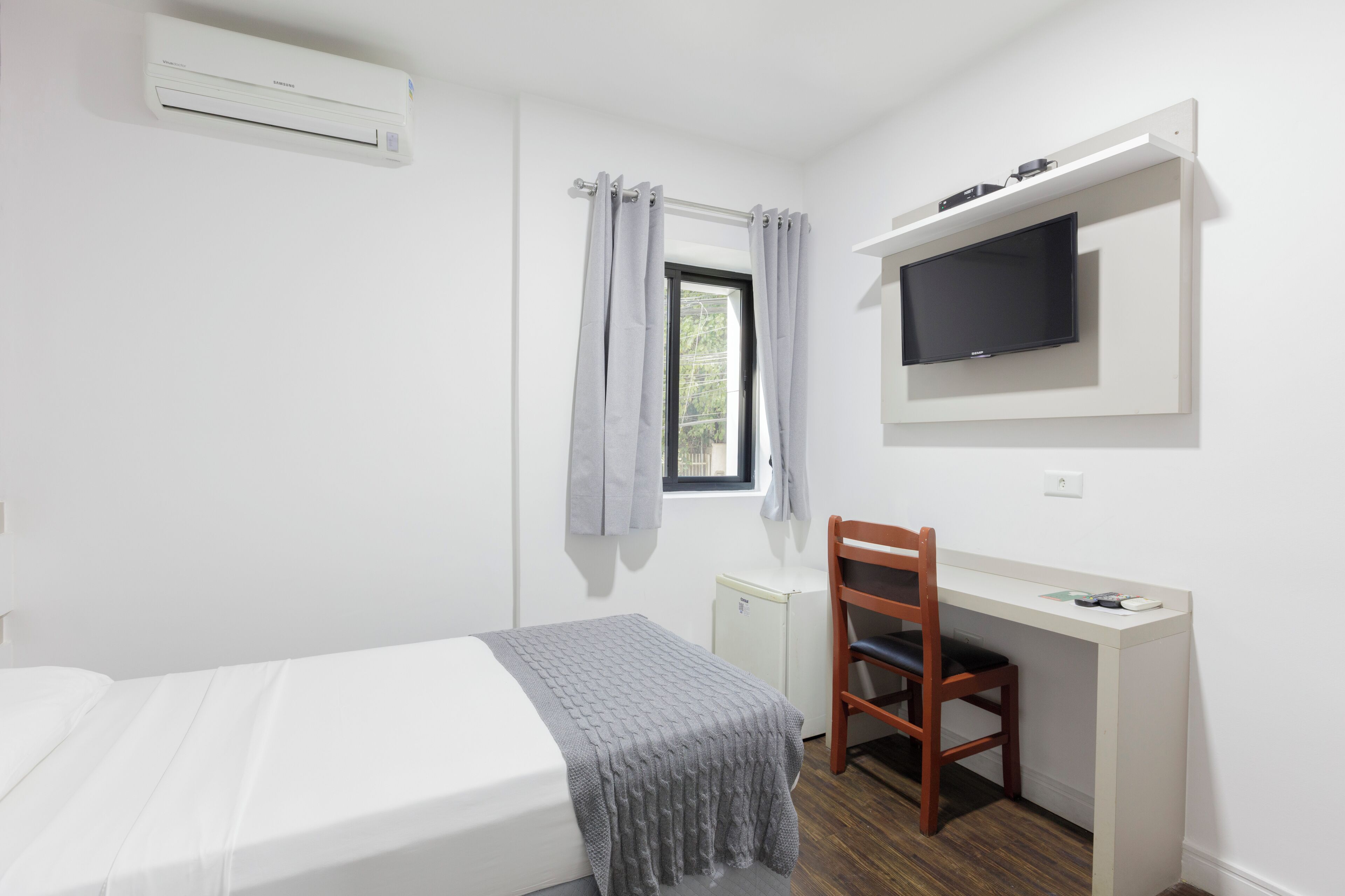 A bedroom with a bed, a desk, a chair, a television, and a window with curtains.