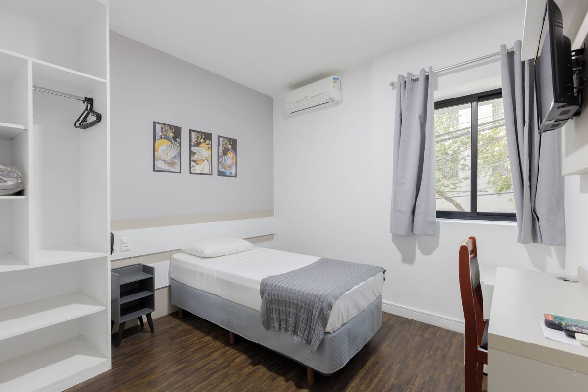A small, neatly organized bedroom with a single bed, a desk, and a chair. There is a window with curtains, an air conditioning unit on the wall, and a shelf with a few items.