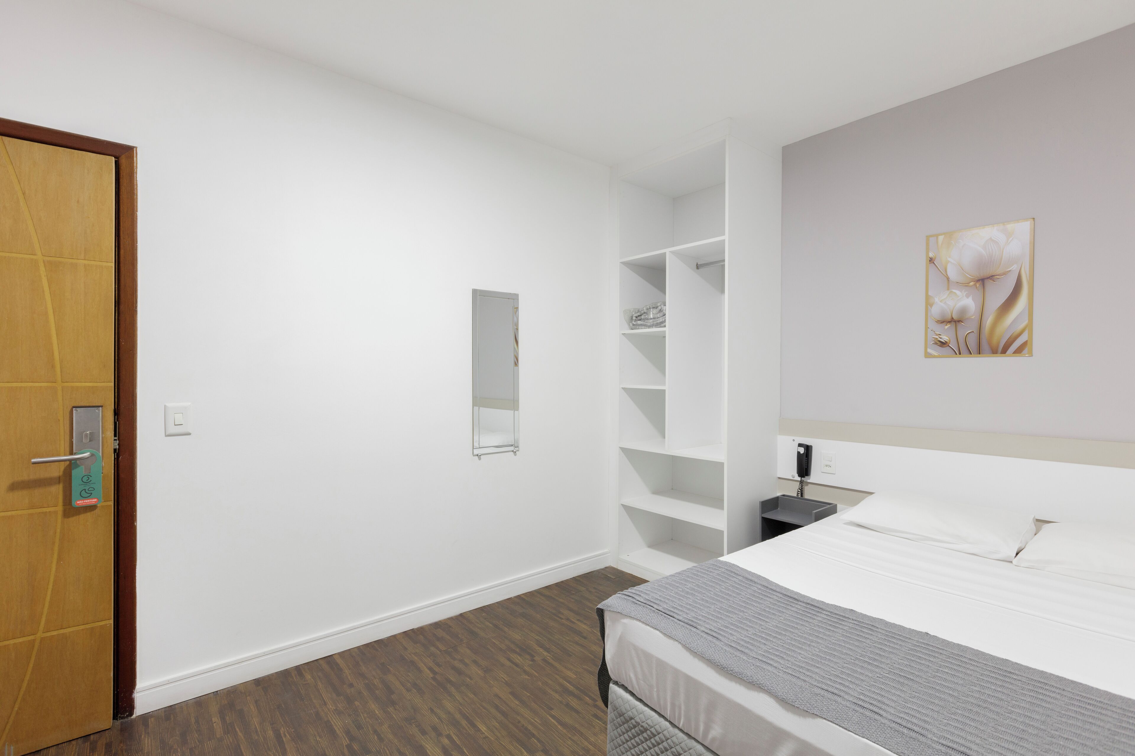 A hotel room with a bed, a wooden door, a white wall, a shelf unit, and a painting.