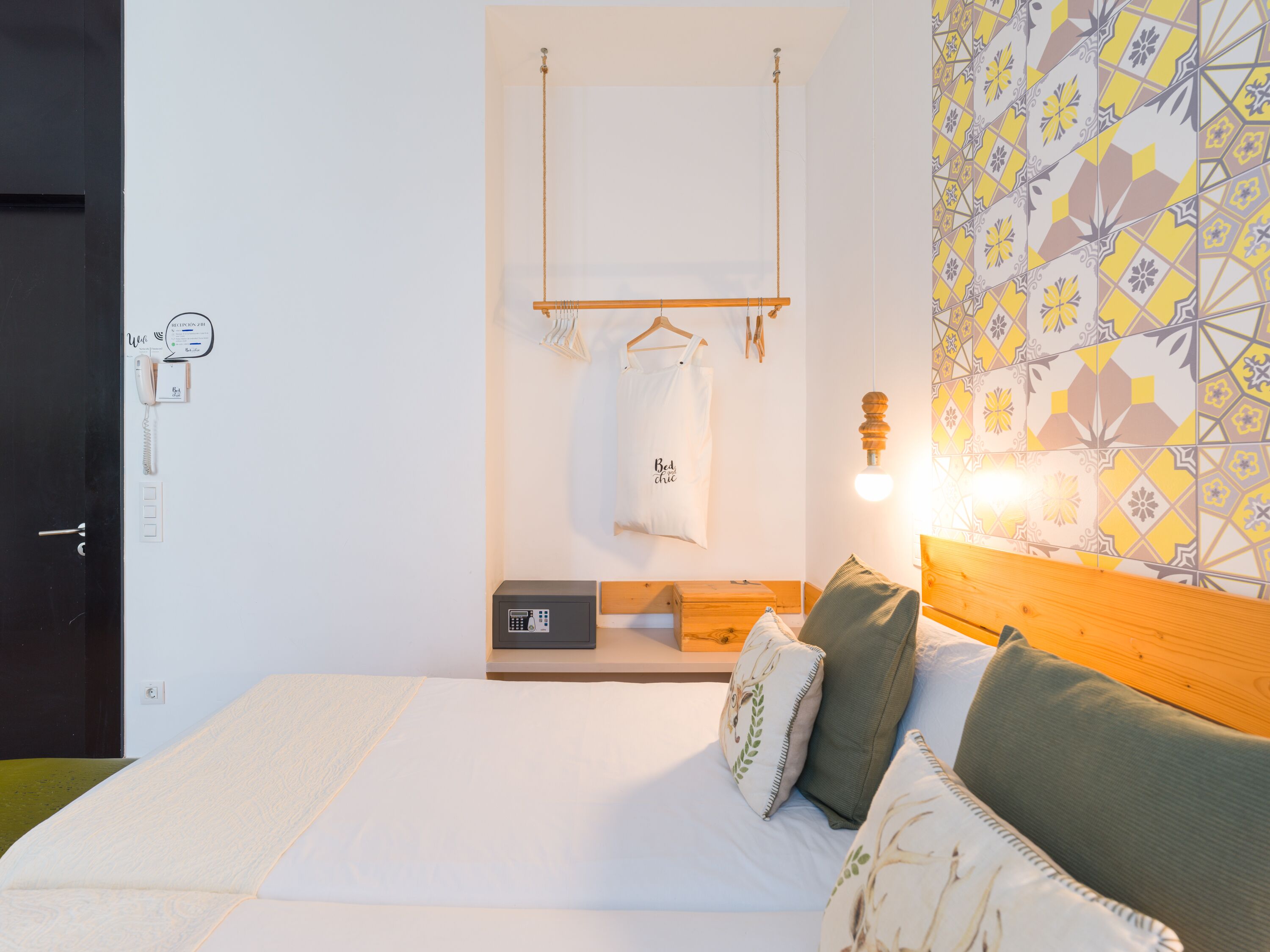 A bedroom with a bed, pillows, a hanging light, a wooden headboard, a bedside table, a wall-mounted telephone, and a patterned wall.