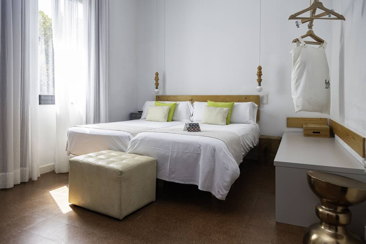 Cama doble con ropa de cama blanca y almohadas verdes, una mesita de noche con lámpara, y una prenda colgada en un perchero de madera.