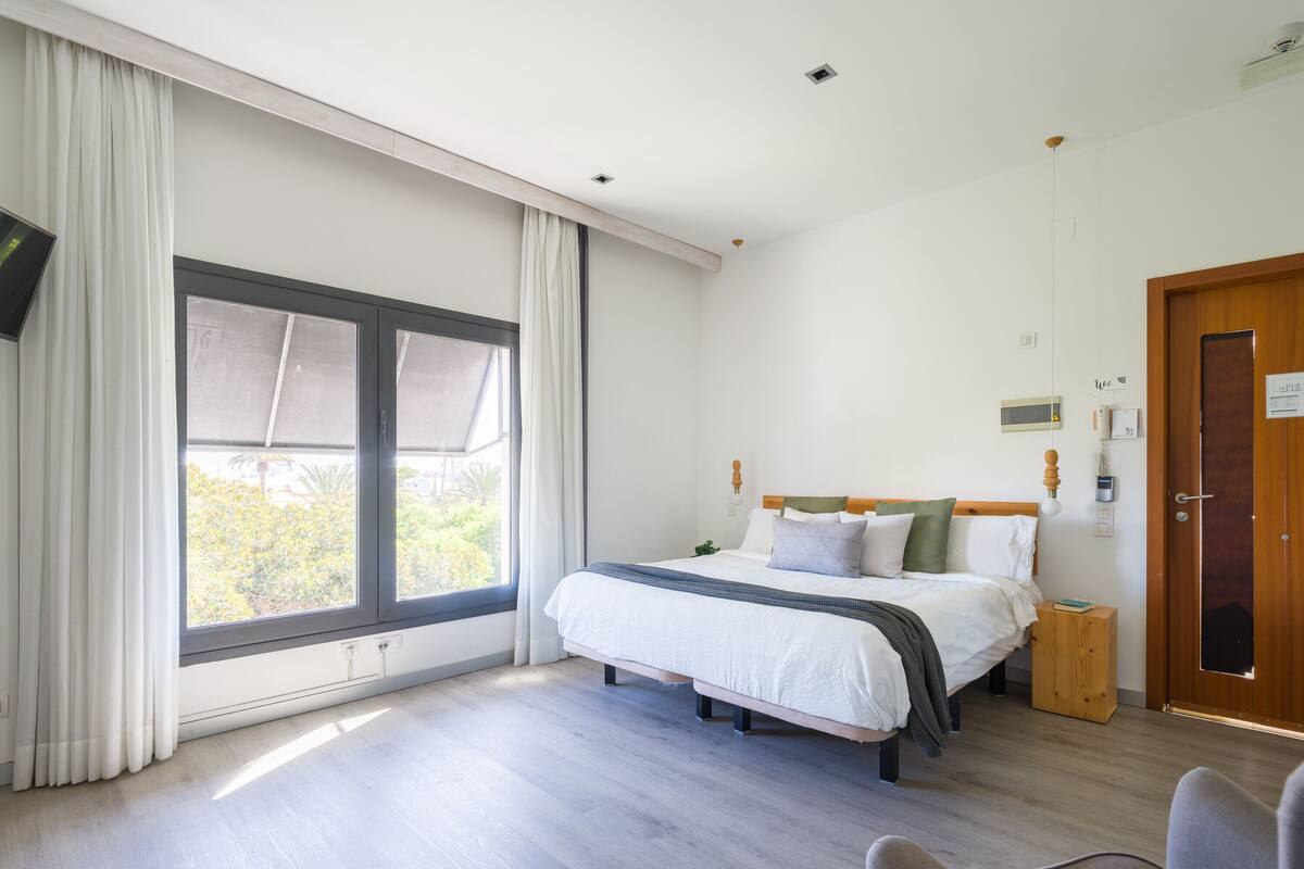Un dormitorio con un ventanal grande, una cama con ropa blanca, una mesita de noche y suelo de madera.