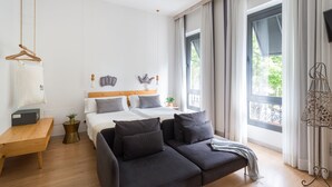Una habitación de hotel moderna con una cama de madera, un sofá oscuro, un banco de madera y ventanas amplias con cortinas.