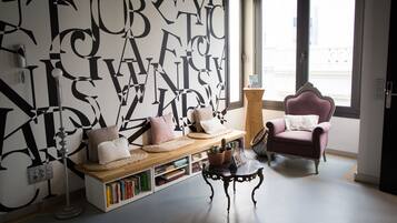 Una sala moderna con un gran mural abstracto en blanco y negro, un sillón rosa, un banco de madera con cojines, una mesita con una planta y una estantería llena de libros.