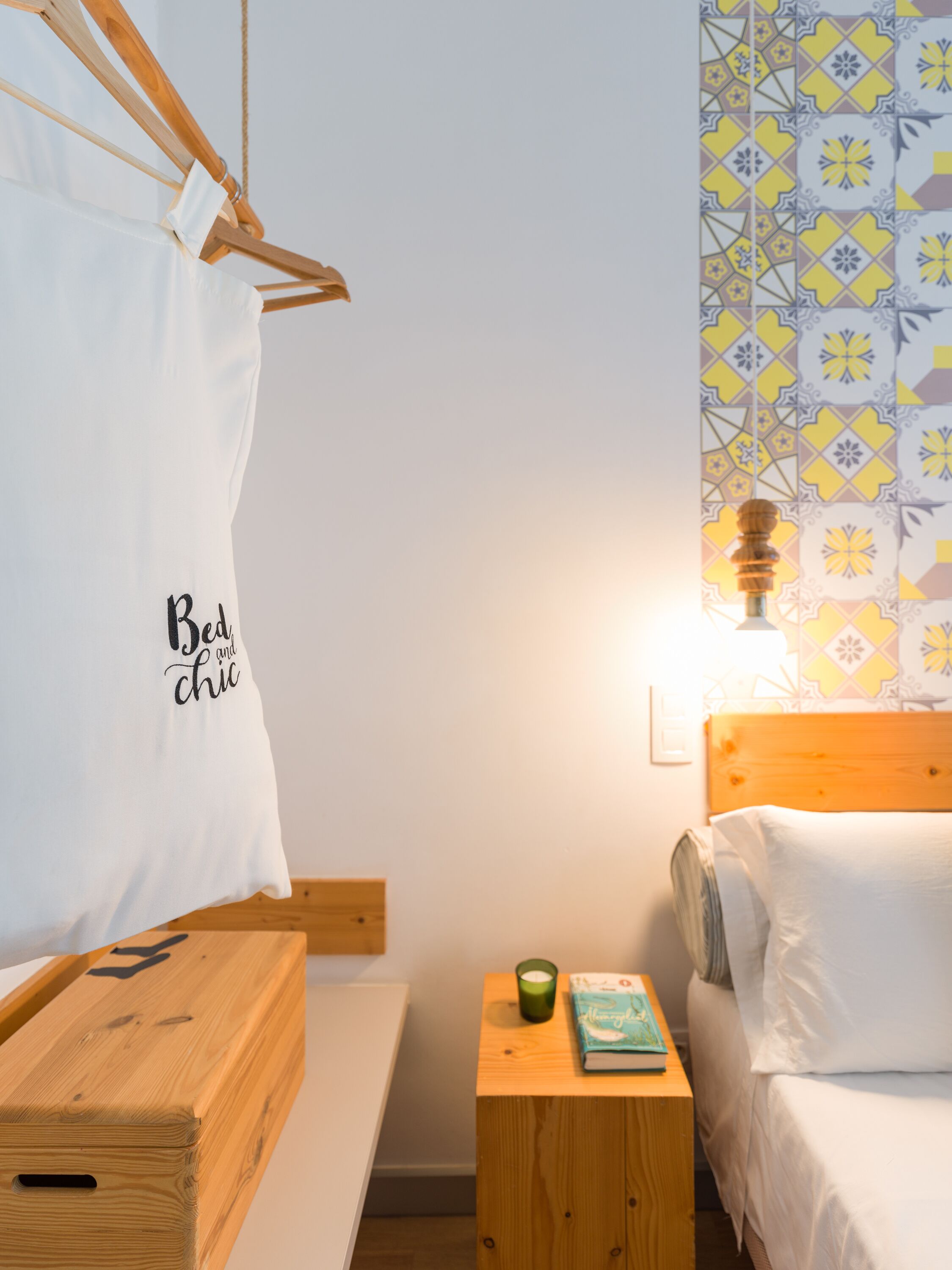 A bedroom with a wooden headboard, a bed with white bedding, a bedside table with a book and a candle, and a hanging white towel with text.