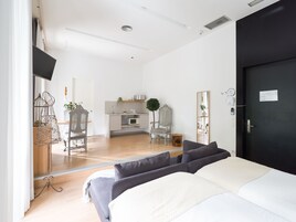 A modern bedroom with a large bed, a dark door, and a kitchenette in the background.