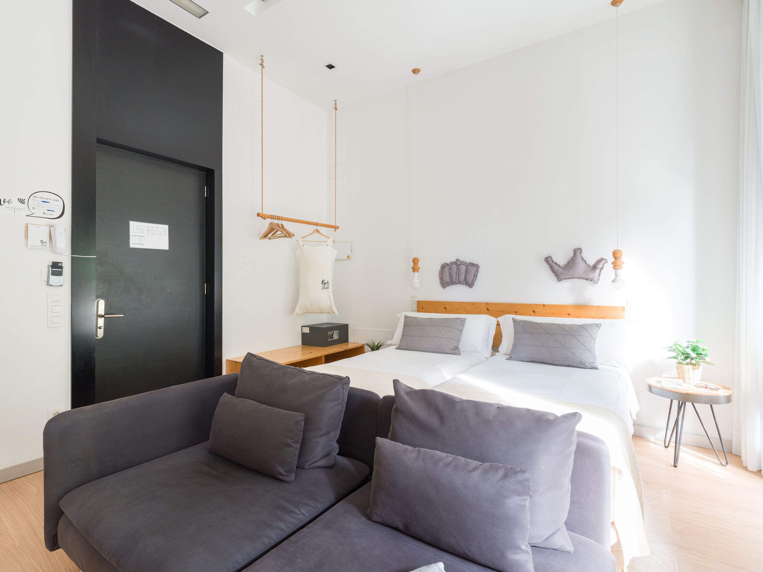 A modern hotel room with a dark door, a sofa with cushions, a bed with two pillows, a bedside table, a small round table with a plant, and a wall-mounted light fixture.