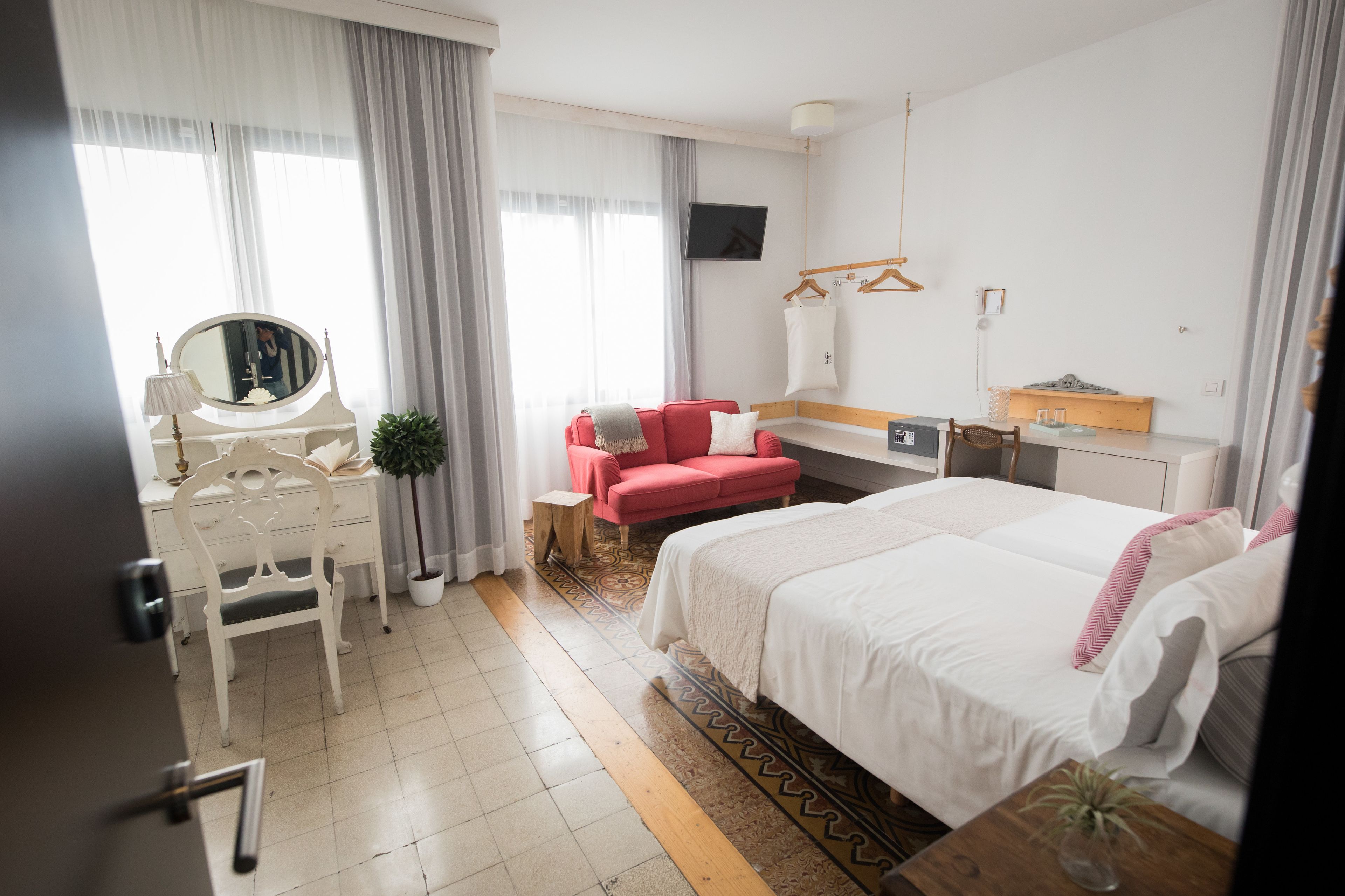Ein Hotelzimmer mit einem Bett, einem Schreibtisch mit Spiegel, einem roten Sessel, einem Fernseher und einem fenster mit Vorhängen.