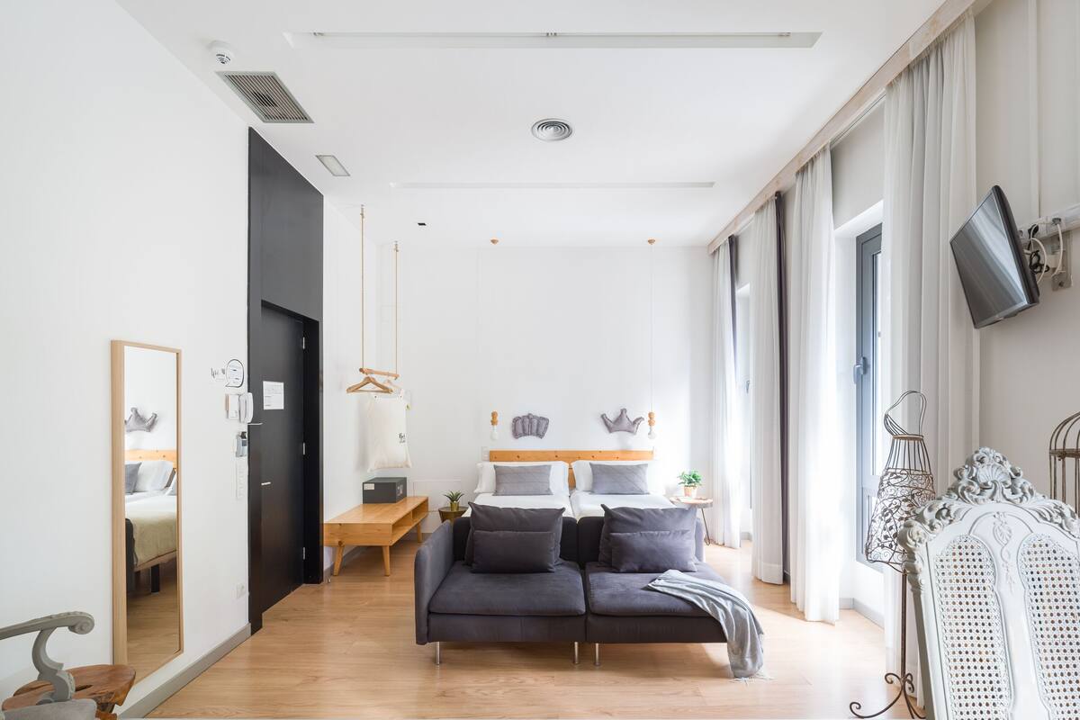 Una habitación de hotel moderna con piso de madera, un sofá gris, una cama con ropa de cama gris, una mesita de noche de madera pequeña y un espejo grande.