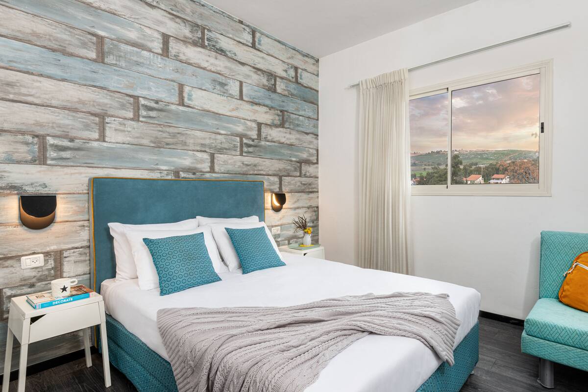 Una cama bien hecha con cabecera azul, una mesita de noche con un libro y una taza, y vista a un paisaje a través de una ventana.