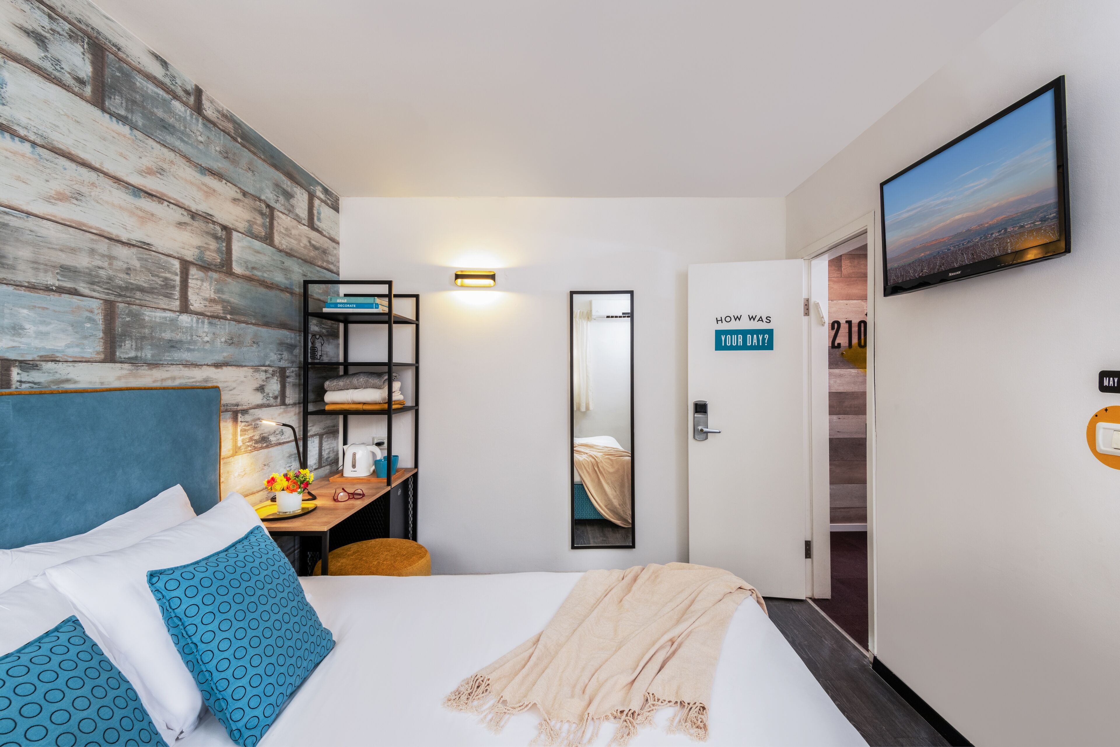 A hotel room with a bed, a desk with a chair, a mirror, a TV, and a door with a sign.