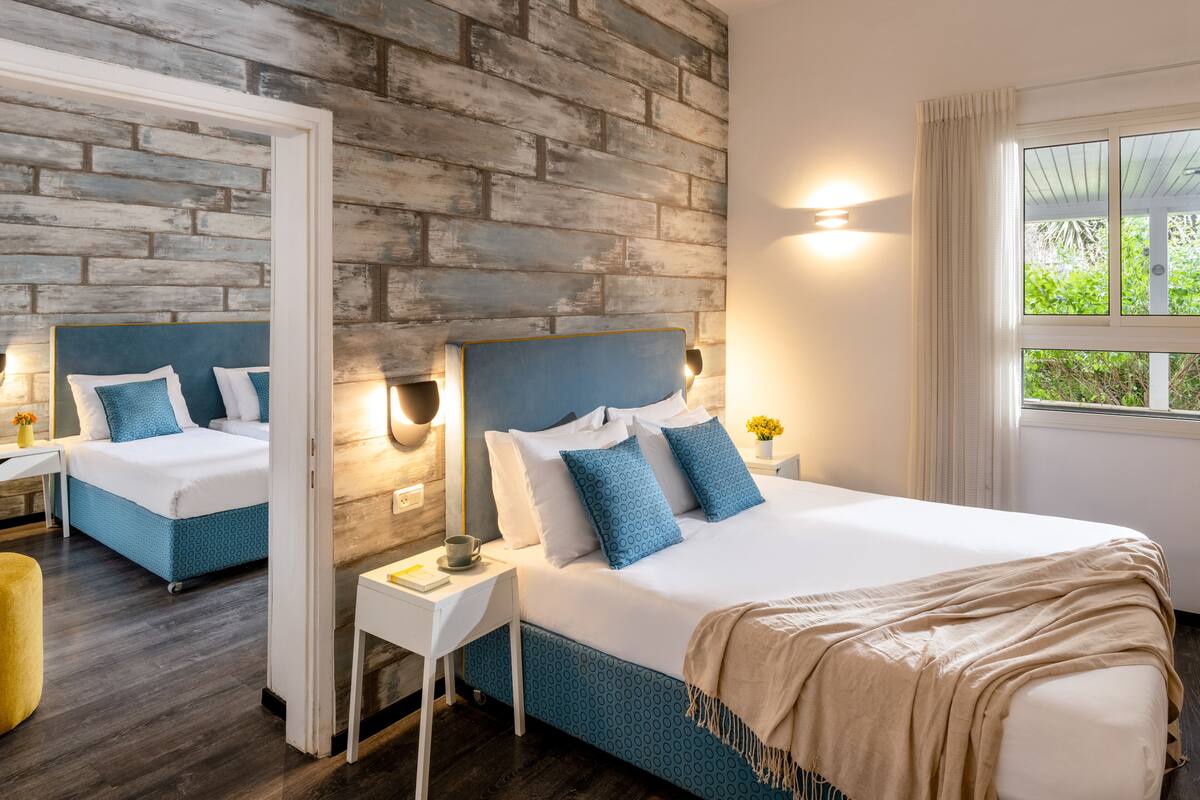 Un dormitorio moderno con pared de ladrillo, una cama grande con ropa de cama azul, una mesita de noche con lámpara y una ventana con cortinas.