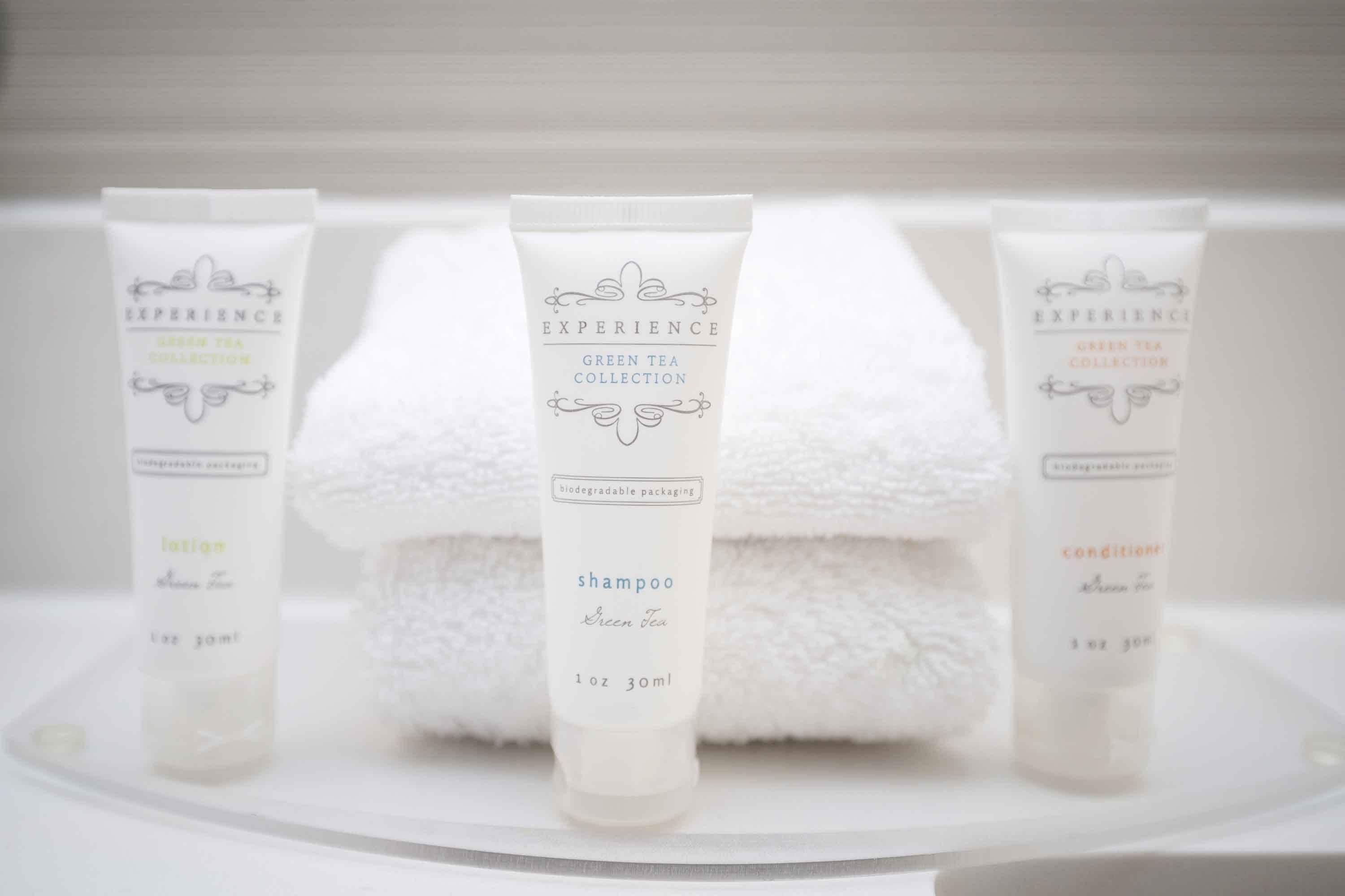 A bathroom with a white towel and three bottles of shampoo labeled "EXPERIENCE GREEN TEA COLLECTION" on a white surface.
