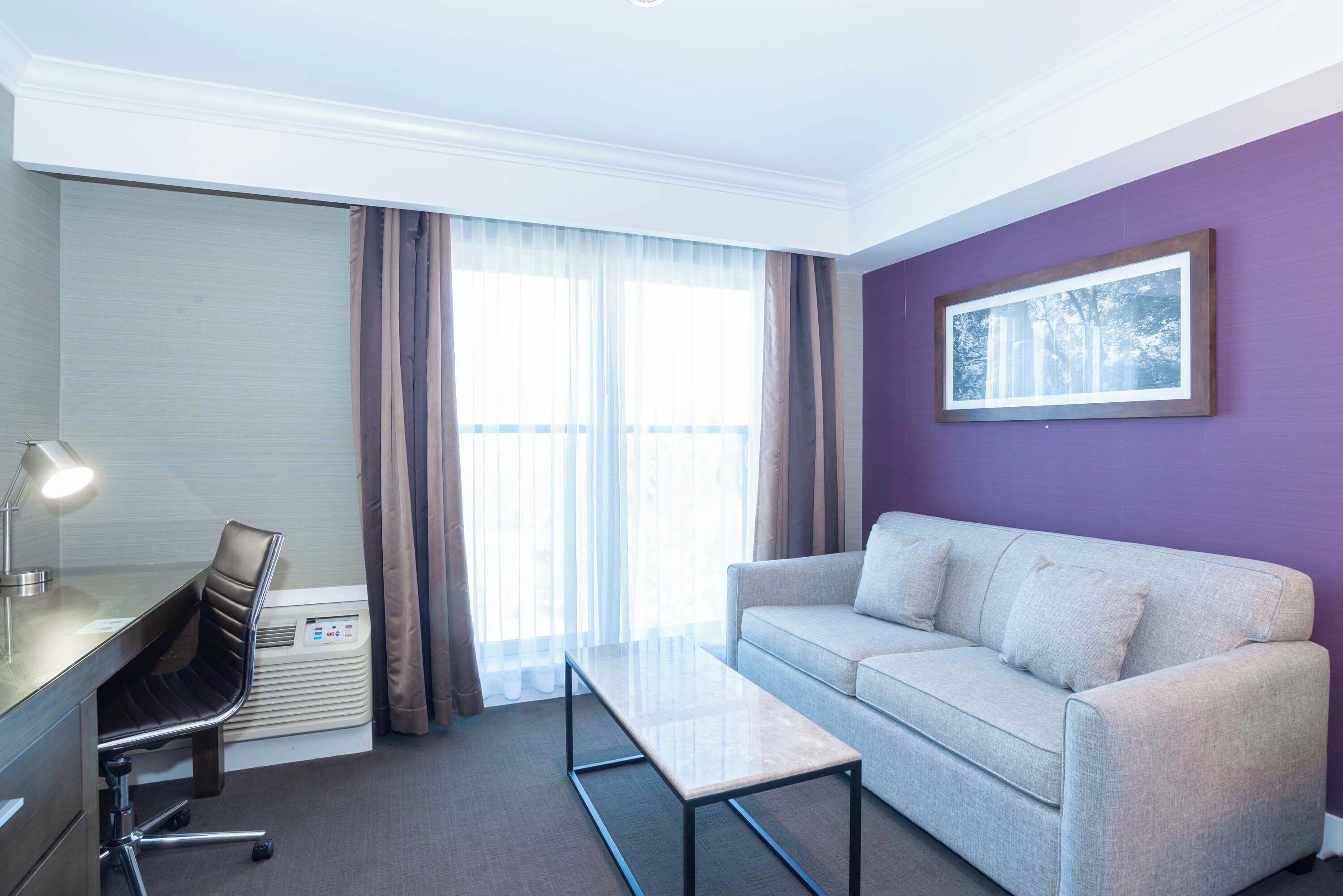 A hotel room with a desk, chair, sofa, and a large window with curtains.