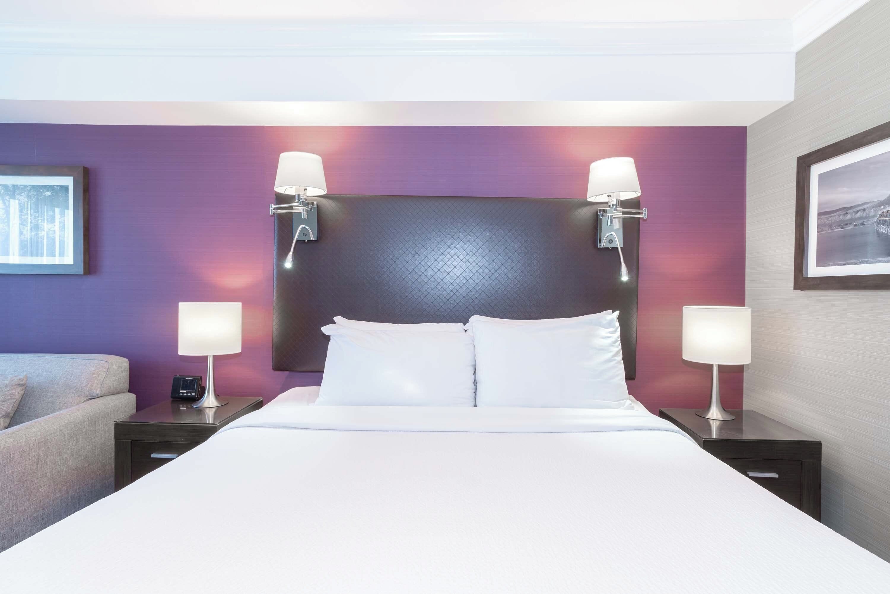 Une chambre d’hôtel avec un grand lit, deux tables de chevet avec des lampes, une tête de lit et un tableau encadré au mur.
