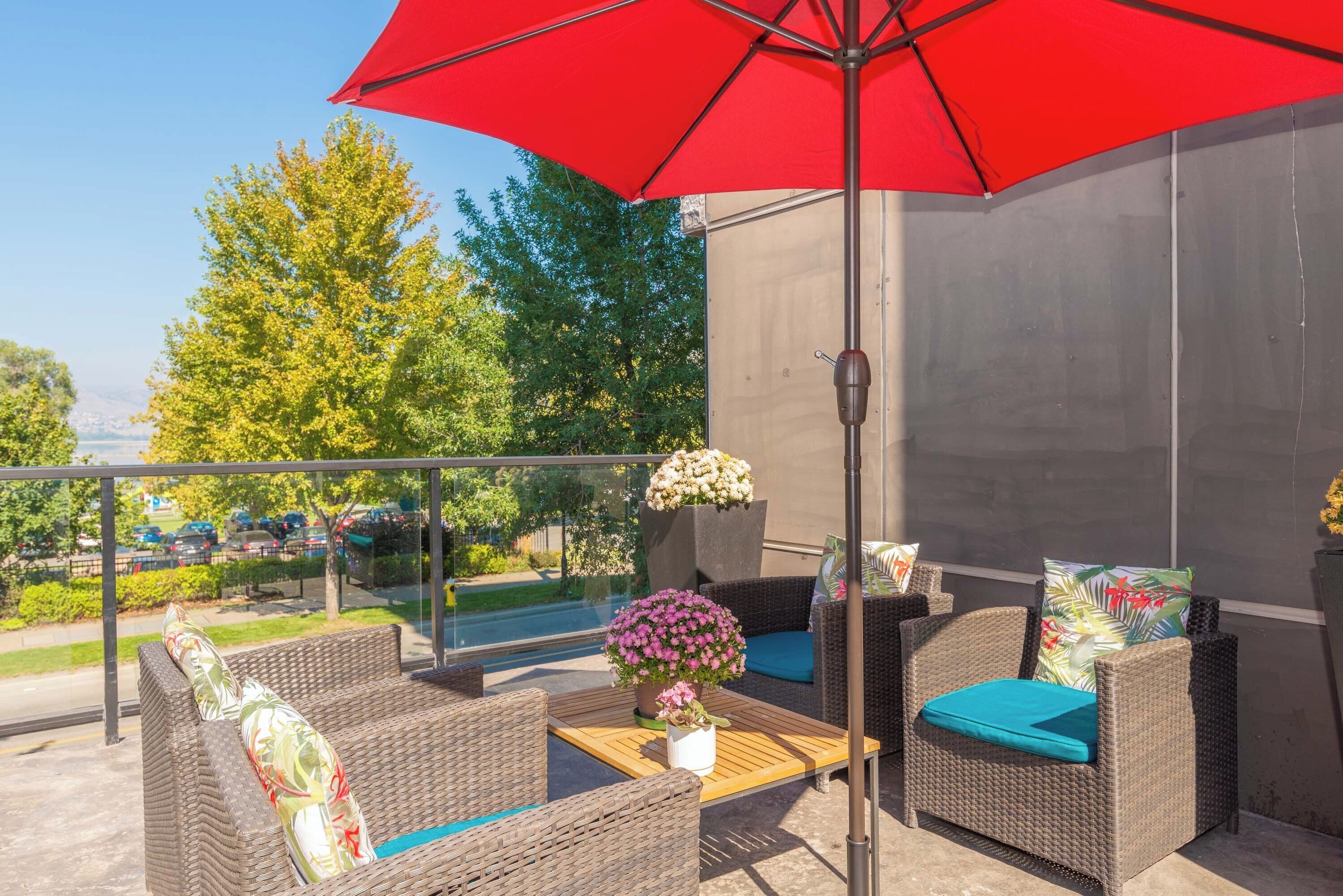 A balcony with wicker furniture, a red umbrella, and a view of trees and parked cars.