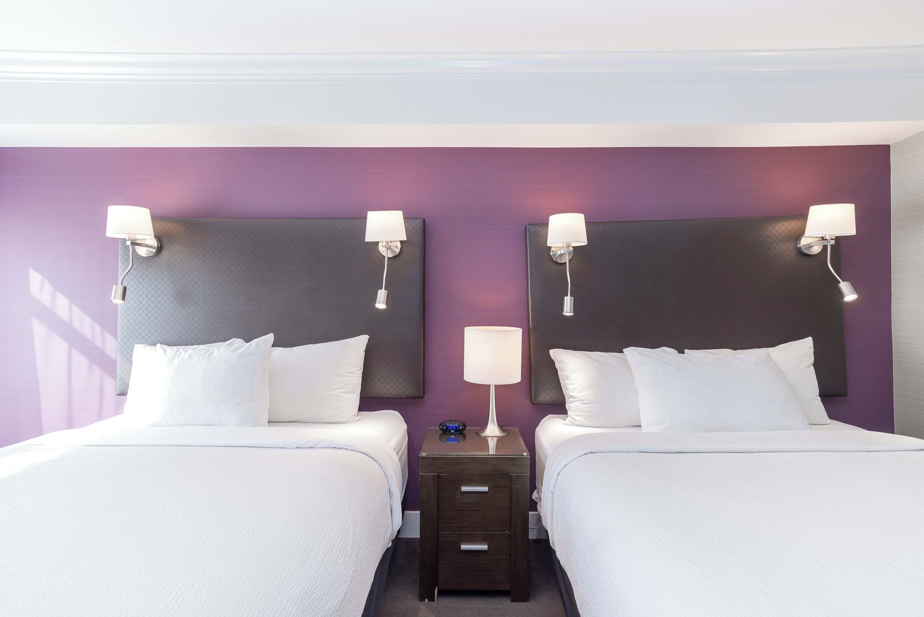 A hotel room with two beds, a nightstand with a lamp, and a wall-mounted clock.