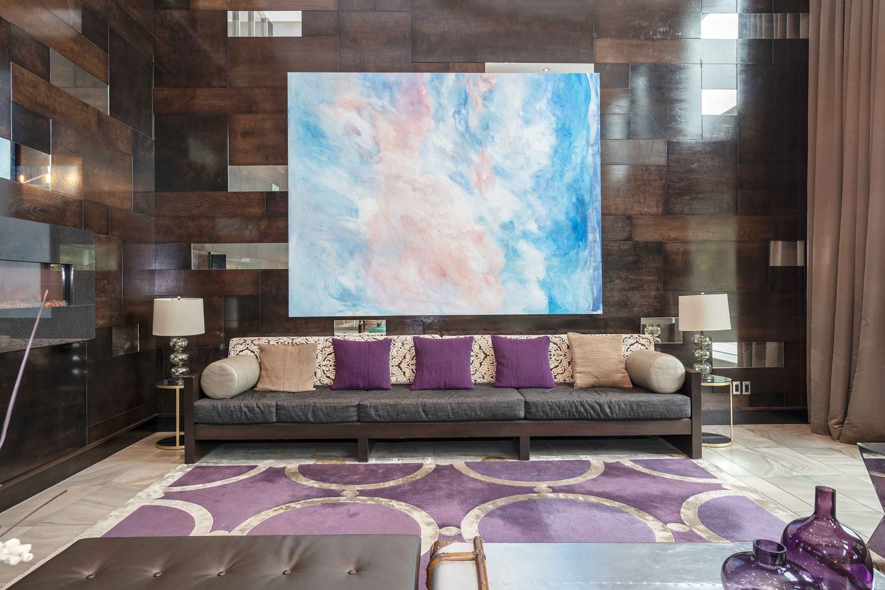 Una sala de estar moderna con un panel de madera oscura, un cuadro abstracto grande, un sofá oscuro con cojines estampados, dos mesitas de noche con lámparas y una alfombra morada con diseño.
