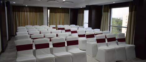 A conference room with rows of white chairs with red sashes, large windows with curtains, and recessed ceiling lights.