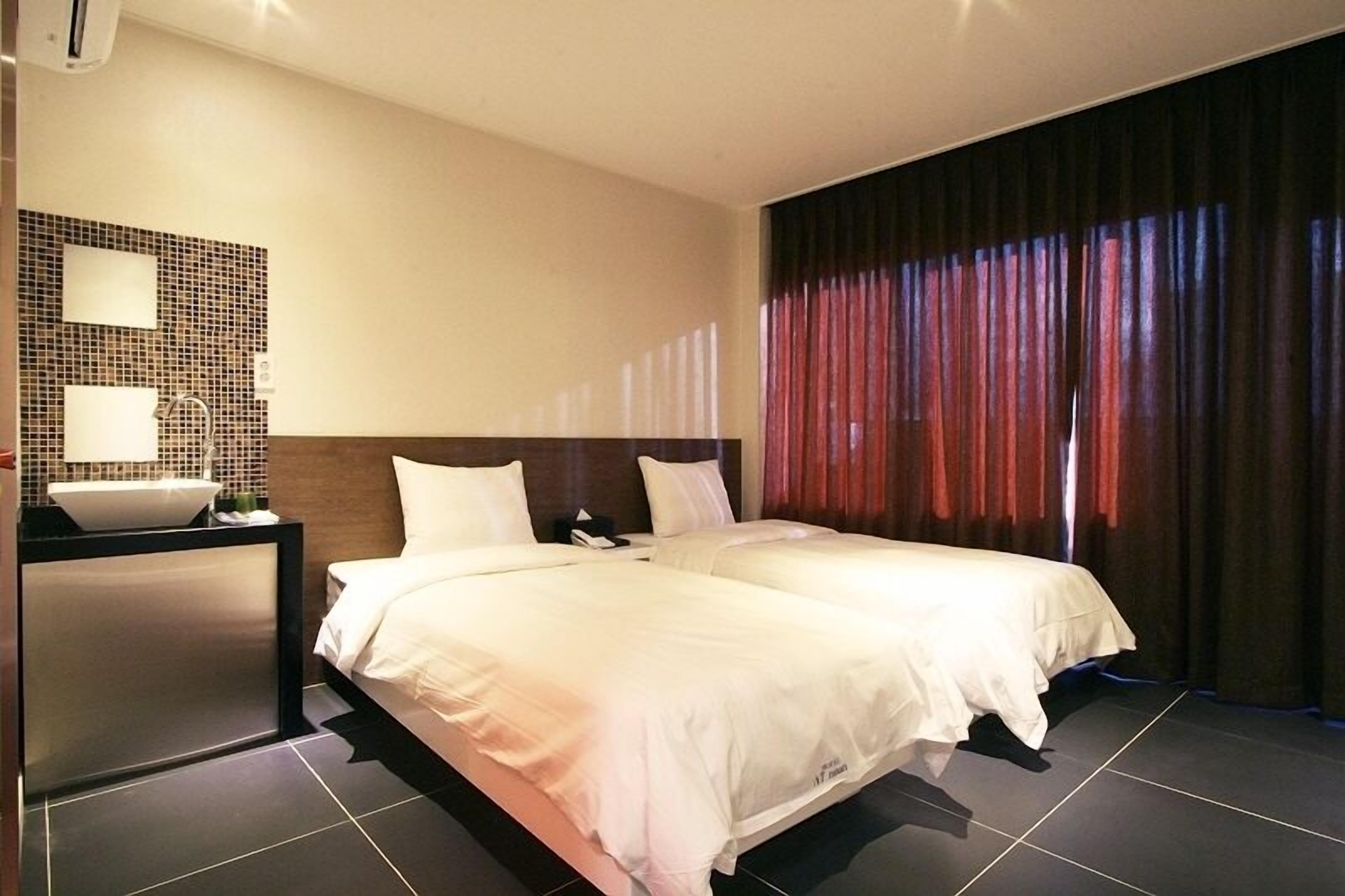 A hotel room with two beds, a bedside table with a lamp, a wall-mounted mirror, and a large window with curtains.