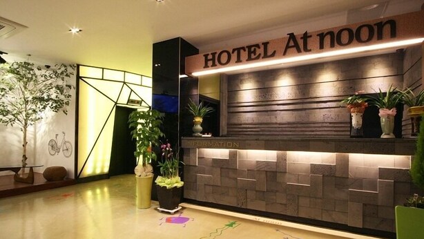 A hotel reception area with a 'HOTEL At noon' sign, an information desk, and decorative plants. - Hotel At noon (Jeju City)