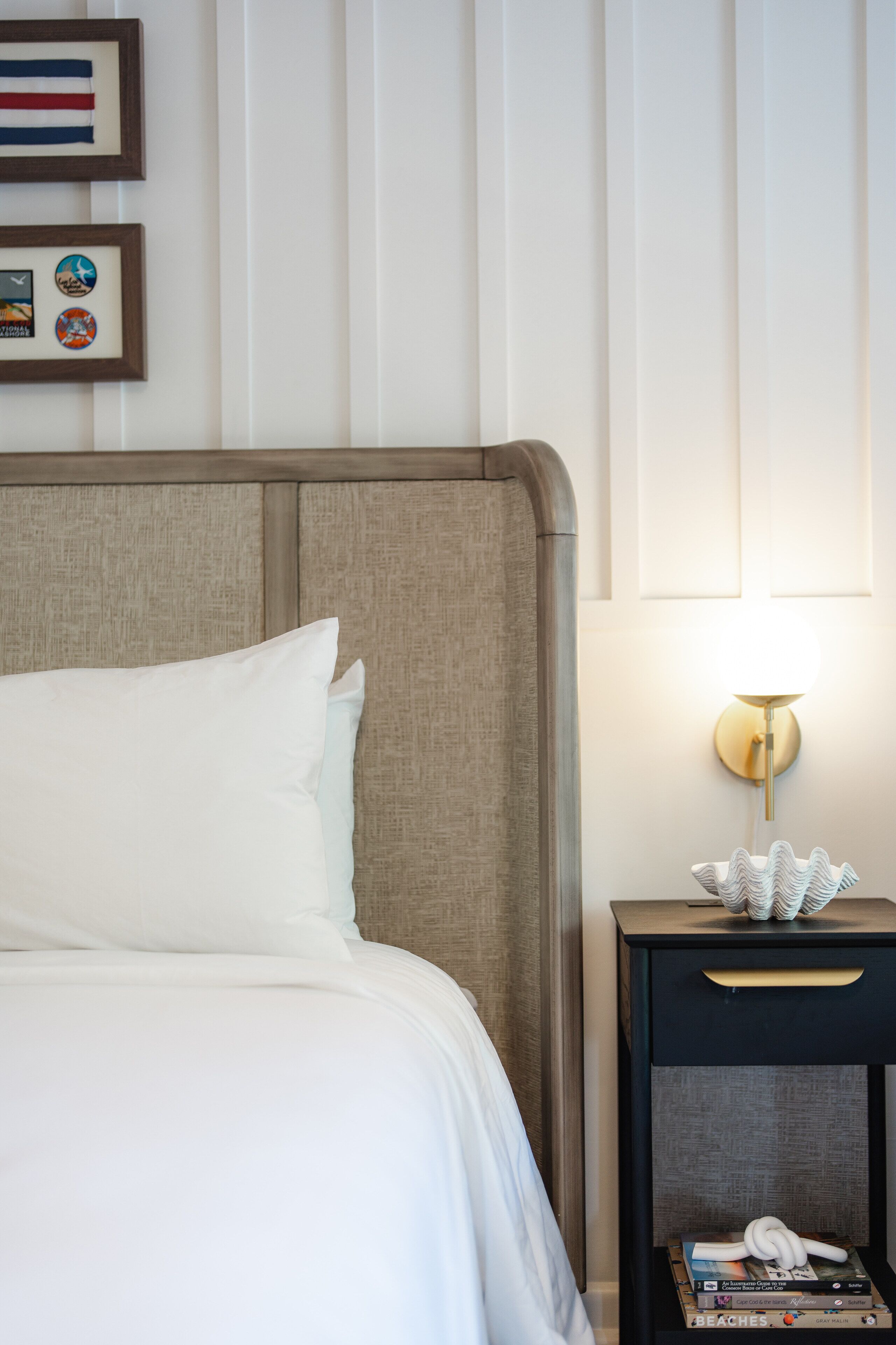 A neatly made bed with white linens, a wooden headboard, a bedside table with a lamp, and framed pictures on the wall.