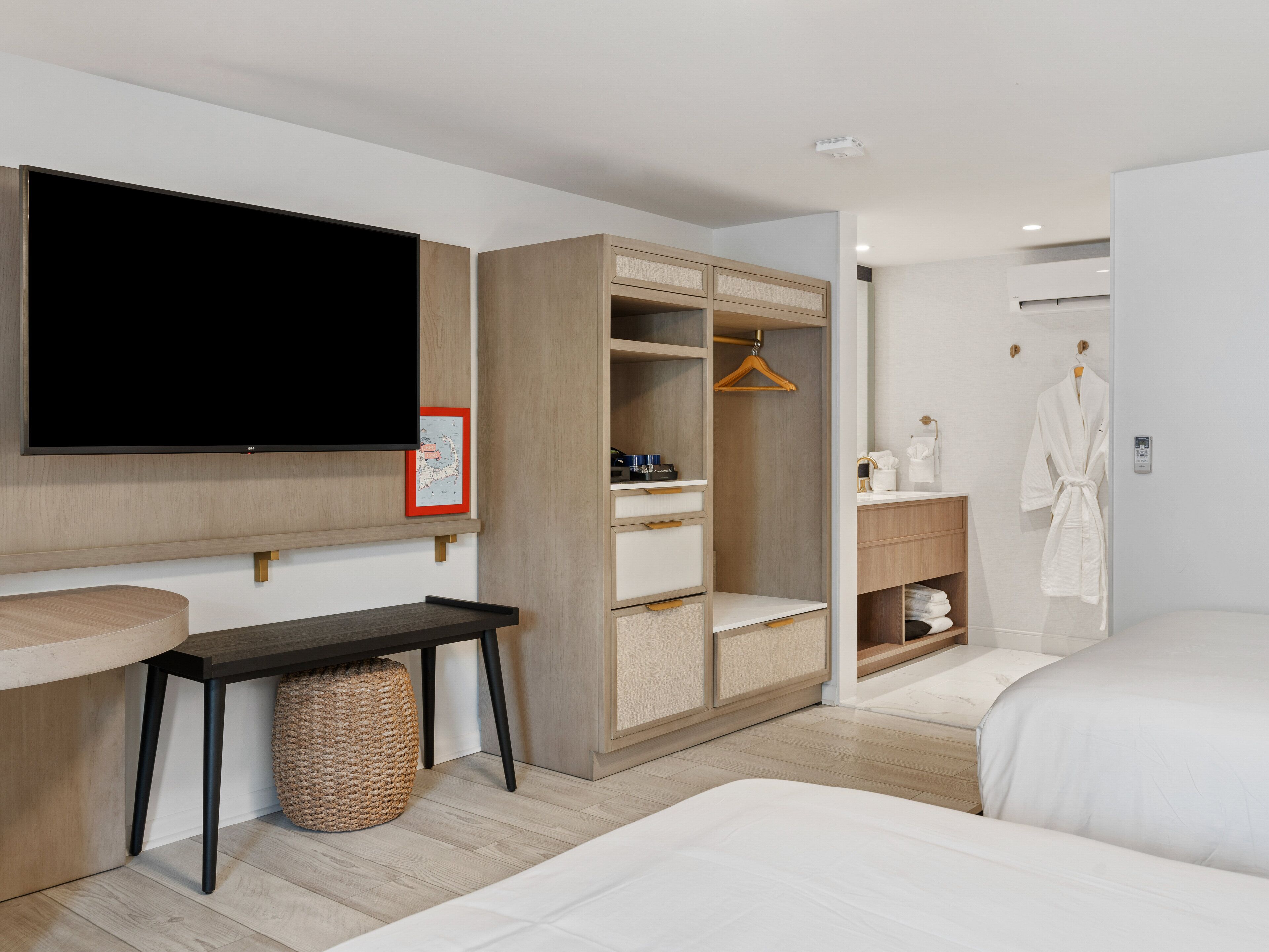 A modern hotel room with a large flat-screen TV, a wooden wardrobe, a bedside table, a hanging robe, and a bathroom with a sink and mirror.